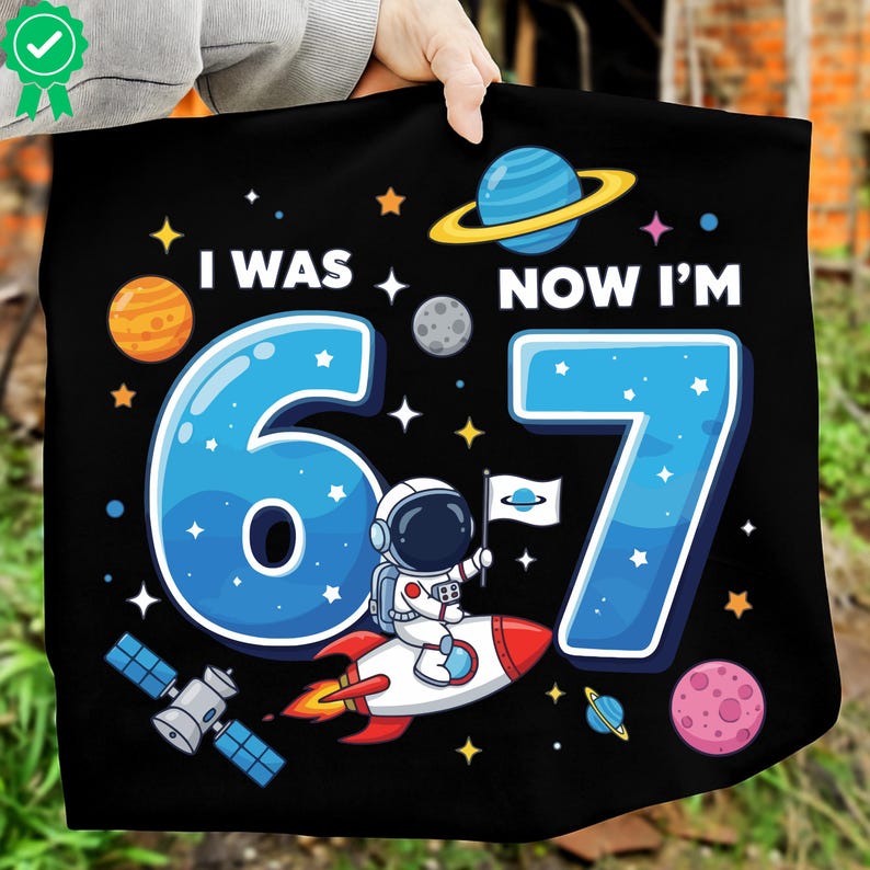 May include: Black textile with a colourful space-themed design. The text reads "I WAS 6, NOW I'M 7" in white and blue. An astronaut on a rocket, planets, stars, and a satellite are also featured.