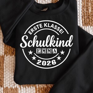 May include: Black crewneck sweatshirt with a white graphic. The graphic reads "ERSTE KLASSE! Schulkind EMMA 2026" in a circular design with stars. The sweatshirt is made of a soft material and is suitable for everyday wear.