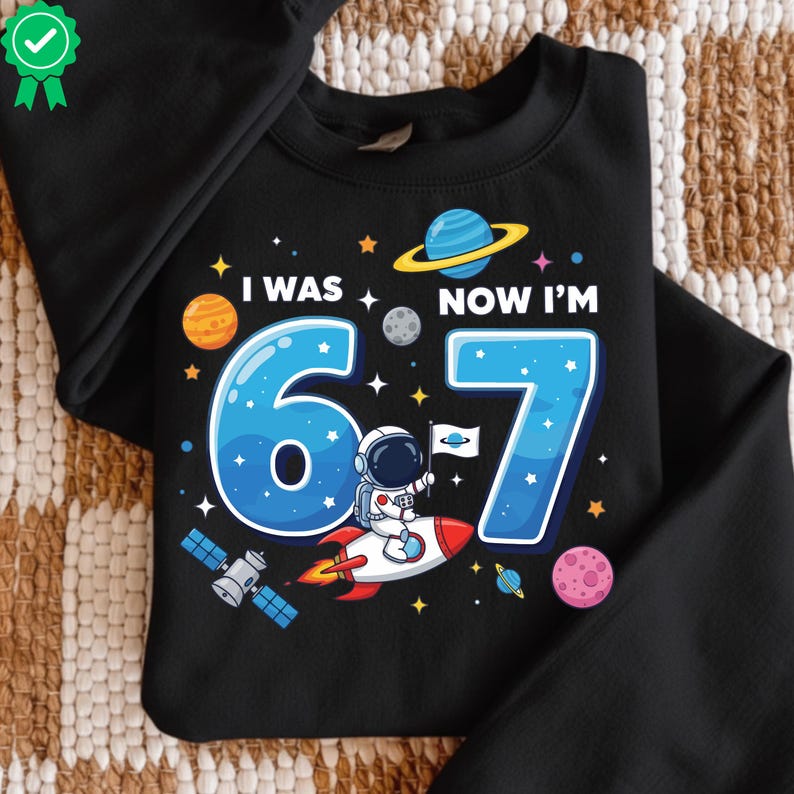 May include: Black sweatshirt featuring a space-themed design. The graphic shows the text "I WAS 6, NOW I'M 7", a blue number 7, an astronaut on a rocket, planets, stars, and a satellite.