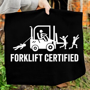 Puede incluir: Bolso tote negro con un gráfico blanco de una carretilla elevadora, una persona siendo arrastrada por la carretilla y dos personas con los brazos levantados. El texto "FORKLIFT CERTIFIED" está impreso debajo.