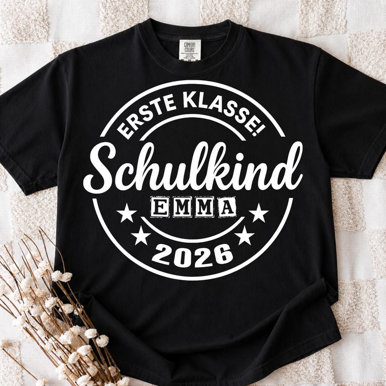 May include: Black t-shirt with a white circular design. The design includes the text "ERSTE KLASSE! Schulkind EMMA 2026" with star accents. The shirt is made of a soft material.