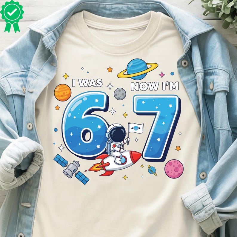 May include: Cream-coloured t-shirt with a space-themed graphic. The design features the text "I WAS 6, NOW I'M 7" in blue, along with planets, stars, and an astronaut on a rocket. A fun birthday shirt.