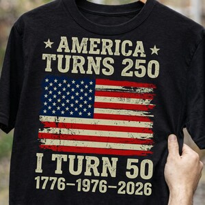 Puede incluir: Camiseta negra con un dise&ntilde;o desgastado de la bandera estadounidense. El texto dice "AMERICA TURNS 250" encima de la bandera y "I TURN 50" debajo, con los a&ntilde;os 1776-1976-2026. El dise&ntilde;o es en blanco, rojo y azul.
