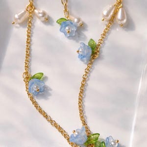 May include: Delicate gold-tone chain necklace with blue flower and pearl accents. The necklace features small, light blue flower pendants with green leaves and faux pearl drops. The design is elegant and feminine, suitable for special occasions or everyday wear.