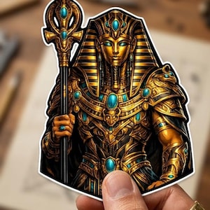 May include: A sticker depicting an Egyptian pharaoh in golden armour, holding a staff. The pharaoh's attire is embellished with turquoise gemstones. The sticker is held by a hand.
