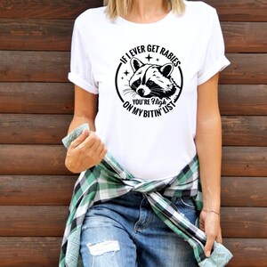May include: White t-shirt with a black graphic of a raccoon and the text "IF I EVER GET RABIES YOU'RE HIGH ON MY BITIN' LIST". The shirt has rolled-up sleeves and is paired with blue jeans.
