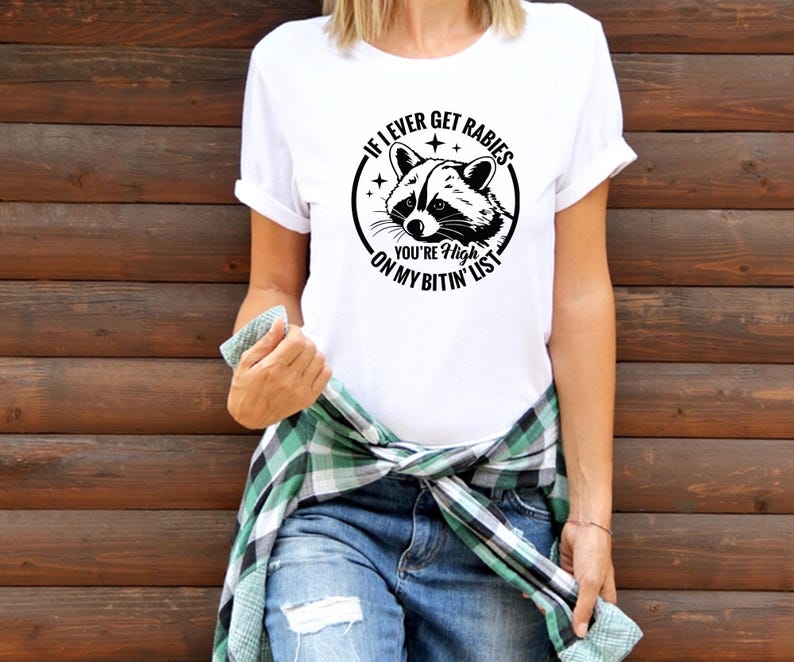 May include: White t-shirt with a black graphic of a raccoon and the text "IF I EVER GET RABIES YOU'RE HIGH ON MY BITIN' LIST". The shirt has rolled-up sleeves and is paired with blue jeans.
