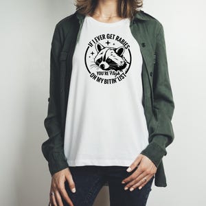 May include: White t-shirt with a black and white raccoon graphic inside a circle, with the text "If I ever get rabies, you're high on my bitin' list". The shirt is a casual style.
