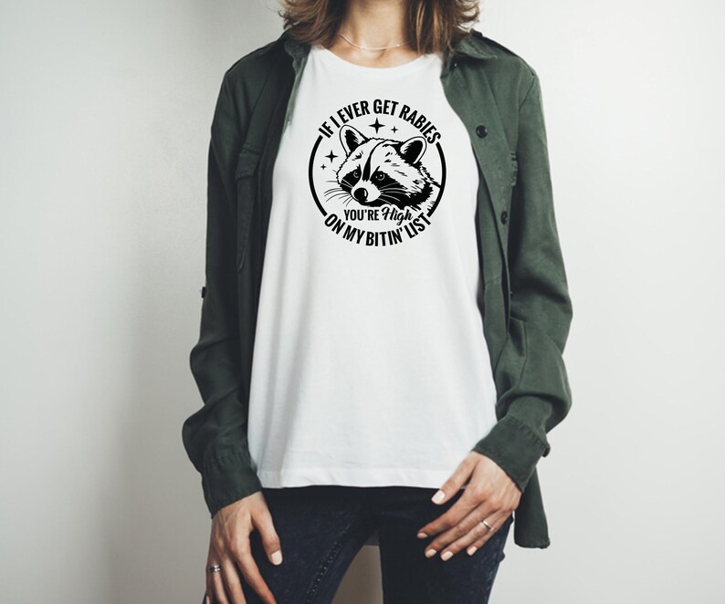 May include: White t-shirt with a black and white raccoon graphic inside a circle, with the text "If I ever get rabies, you're high on my bitin' list". The shirt is a casual style.