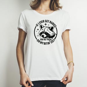 May include: White t-shirt with a black graphic of a raccoon inside a circle, with the text "If I ever get rabies you're high on my bitin' list". The raccoon has a detailed face.