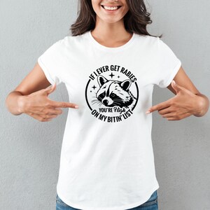 May include: White t-shirt featuring a black and white raccoon graphic inside a circular design. The text reads "If I ever get rabies, you're high on my bitin' list."