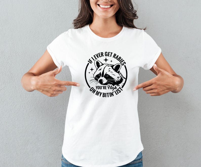 May include: White t-shirt featuring a black and white raccoon graphic inside a circular design. The text reads "If I ever get rabies, you're high on my bitin' list."