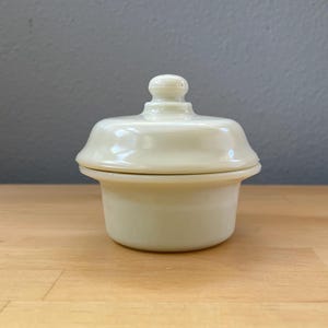 May include: A small, pale cream-colored ceramic lidded jar with a smooth, glossy finish. The container features a cylindrical base and a matching lid with a rounded knob handle centered on top, presenting a simple, uniform, and solid-colored design.