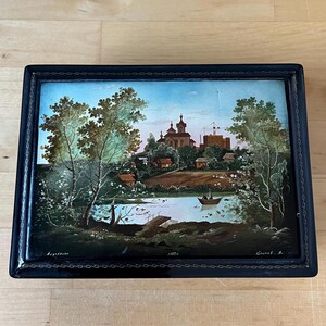 May include: A rectangular black lacquered box with a landscape painting on the lid. The painting depicts a river scene with buildings, trees, and a boat. The artwork is framed by a thin gold border.