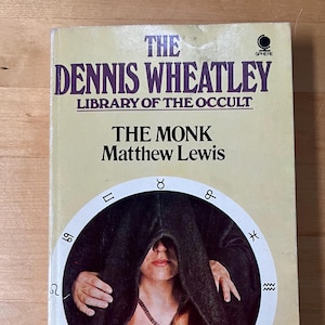 May include: A vintage paperback book titled "The Monk" by Matthew Lewis, part of "The Dennis Wheatley Library of the Occult." The cover features a woman in a dark cloak within a circular design with astrological symbols. The book has a cream-colored background.