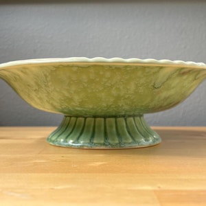 May include: A green ceramic pedestal bowl with a scalloped edge and a textured base. The bowl has a light green glaze with a mottled effect.