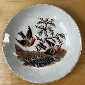 May include: A round, off-white ceramic plate with a detailed bird and foliage design. The illustration features two birds with red accents, perched near a brown and green floral arrangement. The plate has a gold rim.