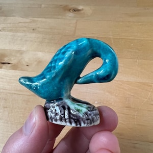May include: A small, teal-colored ceramic swan figurine. The swan has a curved neck and is perched on a textured base with brown, green, and purple hues. The figurine is held in a person's hand.