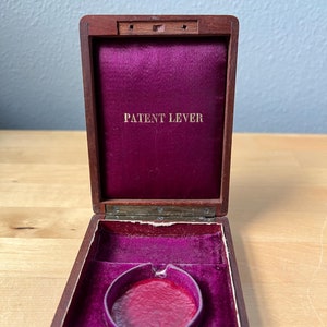 May include: An open wooden box with a red velvet interior. The box has a brass latch and the words "PATENT LEVER" are printed in gold on the inside of the lid.