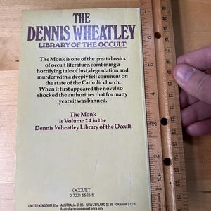 May include: A vintage book titled "The Dennis Wheatley Library of the Occult" with the text "The Monk" visible. The book is Volume 24 and has a beige cover with dark purple text. A wooden ruler is next to the book, measuring approximately 7 inches.