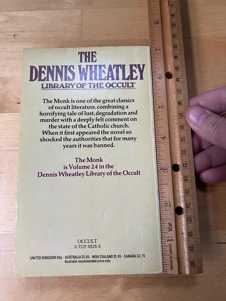 May include: A vintage book titled "The Dennis Wheatley Library of the Occult" with the text "The Monk" visible. The book is Volume 24 and has a beige cover with dark purple text. A wooden ruler is next to the book, measuring approximately 7 inches.