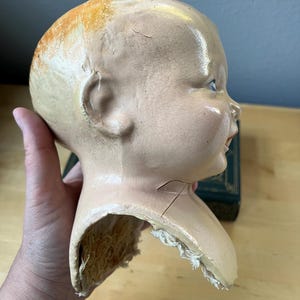 May include: Vintage doll head with a pale complexion, visible cracks, and a partially detached neck. The head has blue eyes, a slight smile, and remnants of orange-toned hair. The doll head is a collectible item.
