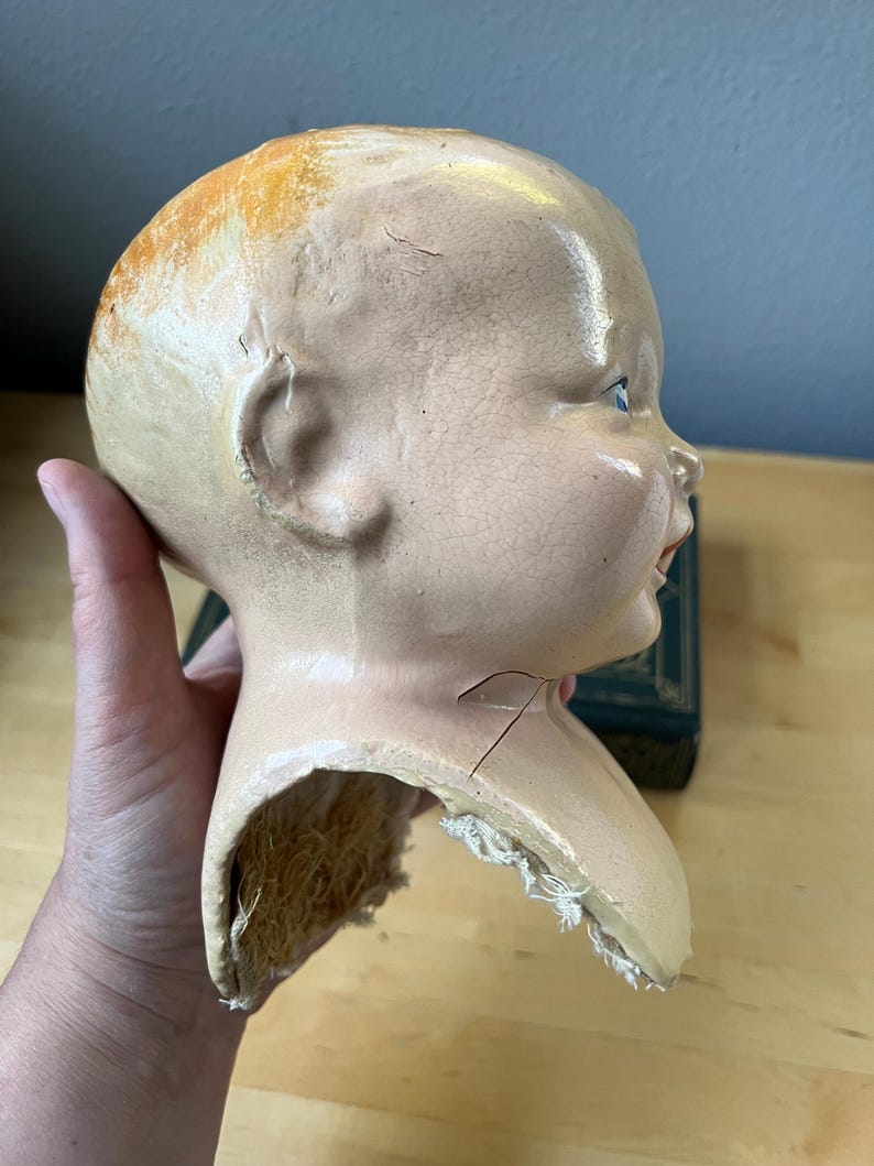 May include: Vintage doll head with a pale complexion, visible cracks, and a partially detached neck. The head has blue eyes, a slight smile, and remnants of orange-toned hair. The doll head is a collectible item.