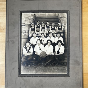 May include: Black and white photo of a high school basketball team posing on the steps of a building. The team is made up of both men and women, and they are all wearing uniforms. The photo is framed in a black and white frame.