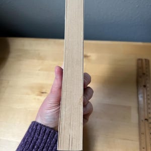 May include: A book with a light tan spine is held by a person wearing a purple sweater. The book's pages are visible, and a wooden ruler is in the background. The book's spine is approximately 1 inch wide.