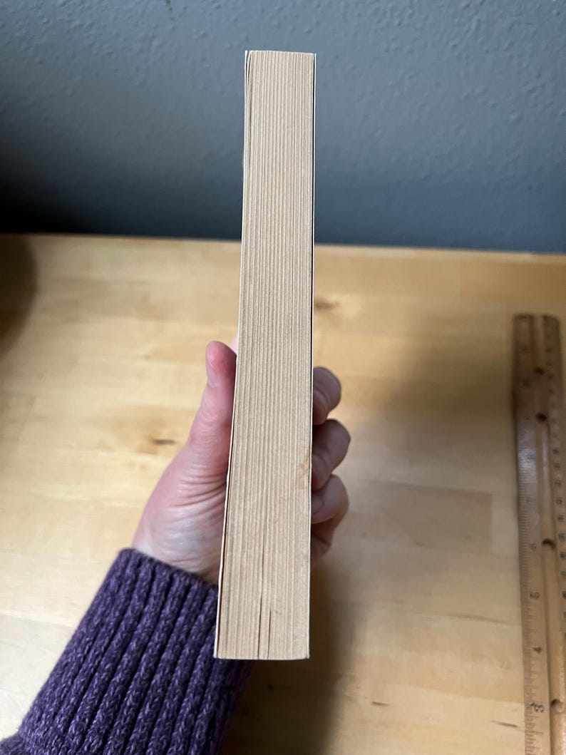 May include: A book with a light tan spine is held by a person wearing a purple sweater. The book's pages are visible, and a wooden ruler is in the background. The book's spine is approximately 1 inch wide.