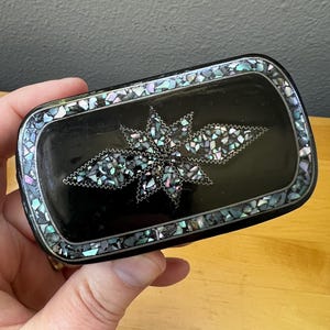May include: A rectangular belt buckle with a smooth black surface, featuring a central star-shaped inlay made of iridescent crushed shell fragments. The border is also decorated with a mosaic of small, multicolored shell pieces.