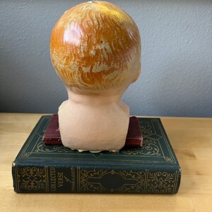 May include: A vintage doll head with painted orange hair, resting on a dark green book with gold lettering. The doll head is peach-colored with visible brushstrokes. The book is titled "Collected Verse" by Edgar A. Guest.
