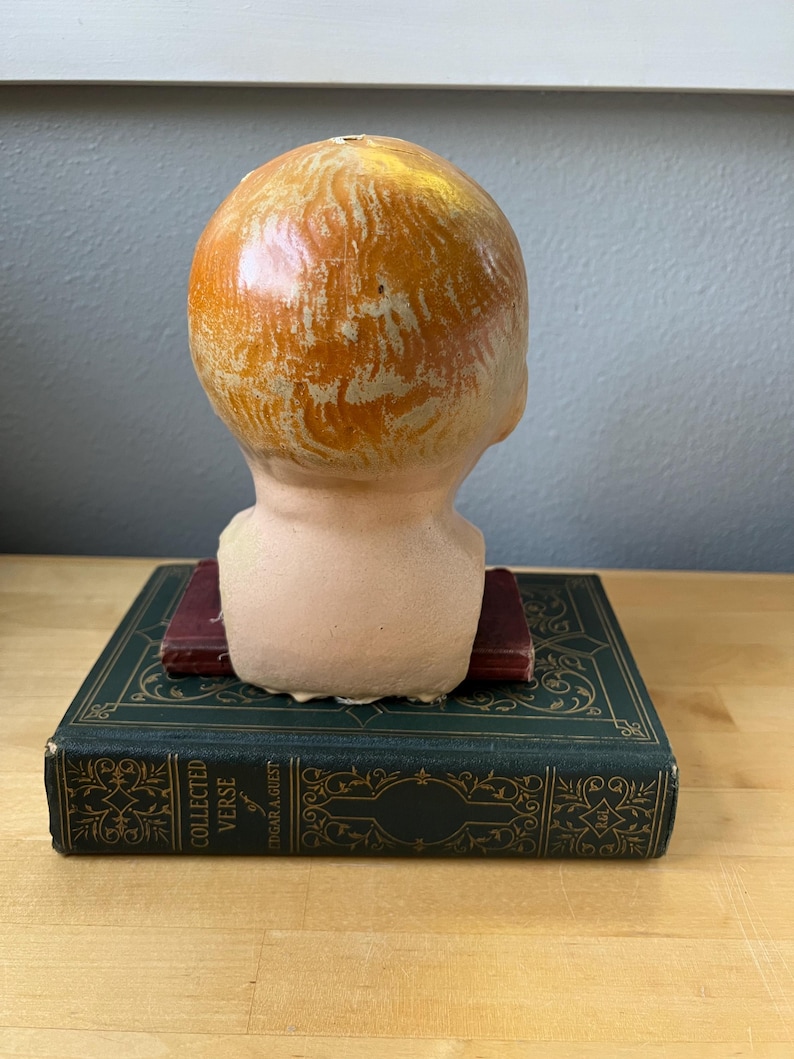 May include: A vintage doll head with painted orange hair, resting on a dark green book with gold lettering. The doll head is peach-colored with visible brushstrokes. The book is titled "Collected Verse" by Edgar A. Guest.