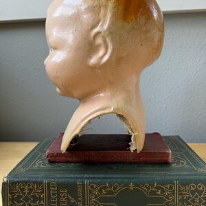 May include: A vintage doll head with a peach complexion and blonde hair, resting on a stack of antique books. The doll head is chipped at the neck. The books are dark green and maroon.