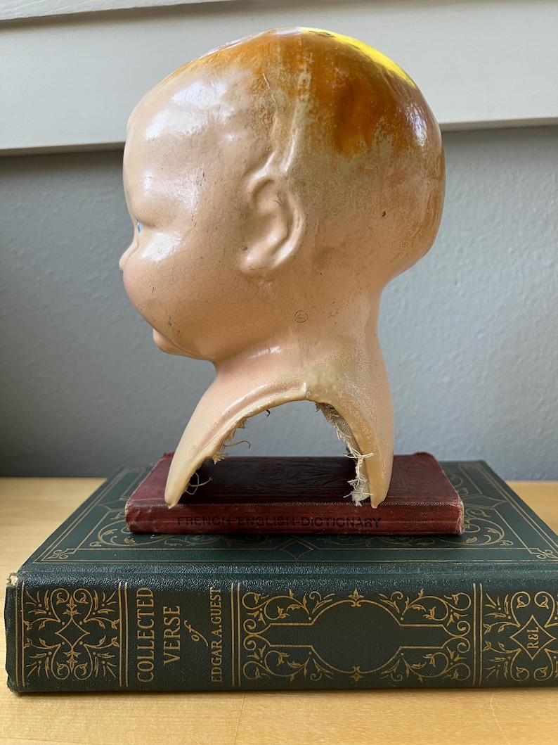 May include: A vintage doll head with a peach complexion and blonde hair, resting on a stack of antique books. The doll head is chipped at the neck. The books are dark green and maroon.