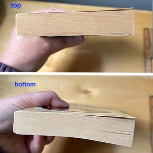 May include: A used book with a tan cover and aged pages. The top and bottom edges of the book are visible, showing the thickness of the pages. The book is held in a person's hand.