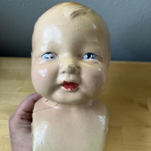 May include: A vintage baby doll head with blue eyes and red lips. The doll head is painted in a flesh tone with visible cracks in the paint. The doll's head is attached to a square-shaped torso.