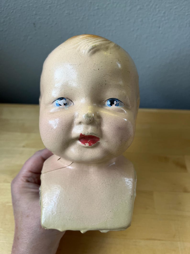 May include: A vintage baby doll head with blue eyes and red lips. The doll head is painted in a flesh tone with visible cracks in the paint. The doll's head is attached to a square-shaped torso.