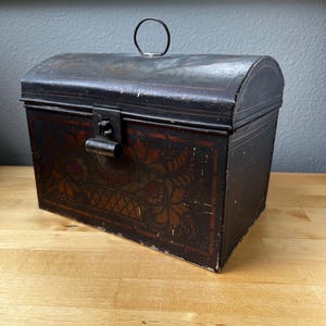 May include: A vintage black metal trunk with a curved top and a handle. The trunk features ornate floral designs in shades of brown and red. A metal latch secures the front. The trunk sits on a wooden surface.