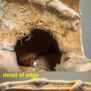 May include: Close-up of a textured, brown, and beige art piece. The artwork features a rough, organic aesthetic with visible layers of fabric and a dark interior. The edges are detailed with a light-colored, glossy material.
