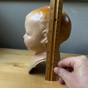 May include: A vintage doll head, painted in flesh tones with a brown-orange hair. The head is approximately 7 inches tall. The doll head is on a wooden surface.