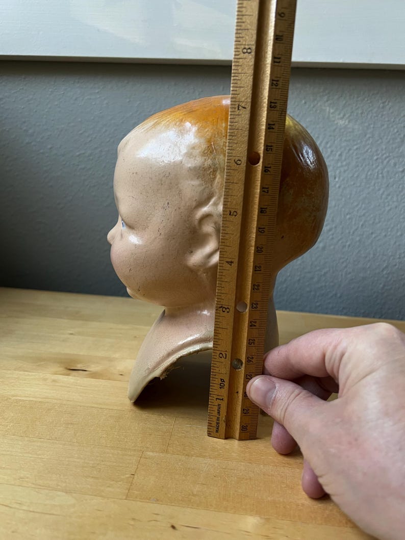 May include: A vintage doll head, painted in flesh tones with a brown-orange hair. The head is approximately 7 inches tall. The doll head is on a wooden surface.