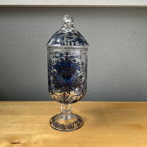 May include: A clear glass jar with a lid, featuring a blue and gold floral design. The jar has a pedestal base.
