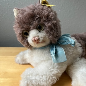 May include: A vintage plush cat toy with gray and white fur, brown eyes, and a blue ribbon around its neck. The cat has a yellow tag on its ear.