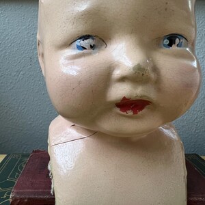 May include: An antique doll head with a cracked, aged appearance. The doll head has blue eyes, red lips, and a pale pink complexion. The head is resting on a dark green and maroon book.