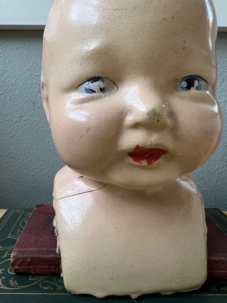May include: An antique doll head with a cracked, aged appearance. The doll head has blue eyes, red lips, and a pale pink complexion. The head is resting on a dark green and maroon book.