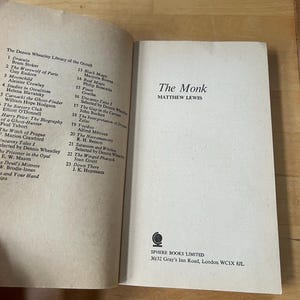 May include: An open book titled "The Monk" by Matthew Lewis, part of "The Dennis Wheatley Library of the Occult." The book's cover is white with black text. A list of other books is visible on the left page.