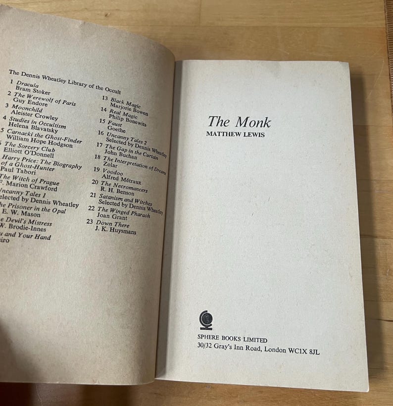 May include: An open book titled "The Monk" by Matthew Lewis, part of "The Dennis Wheatley Library of the Occult." The book's cover is white with black text. A list of other books is visible on the left page.