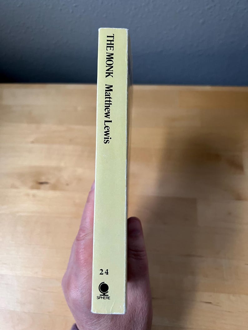 May include: A book spine with the title "THE MONK" by Matthew Lewis. The spine is a light yellow color with the number "24" and the "SPHERE" logo at the bottom. The book is held by a hand.