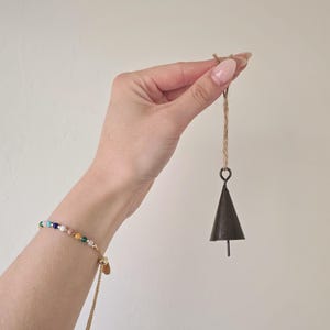 May include: A small, dark metal bell with a triangular shape hangs from a natural twine cord. A beaded bracelet with colourful stones and a gold chain is visible on a wrist.